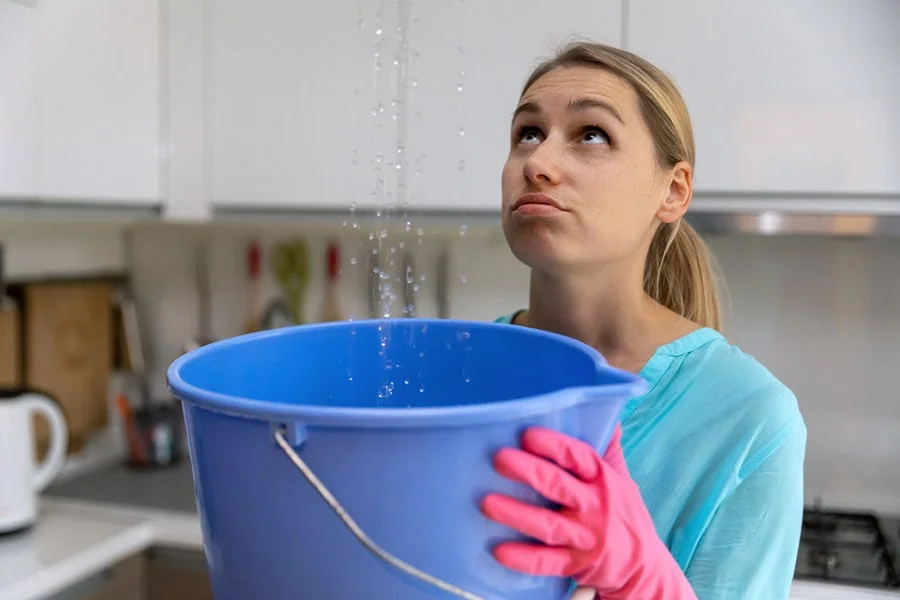 A concerned woman in a kitchen holding a large blue plastic bucket to catch water leaking from the ceiling.